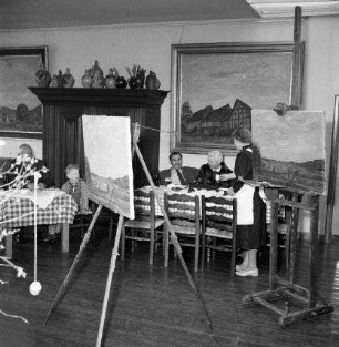 Ateliergasthaus. Gaststube. Ansicht mit Gästen und Kellnerin, im Vordergrund Staffeleien mit Gemälden