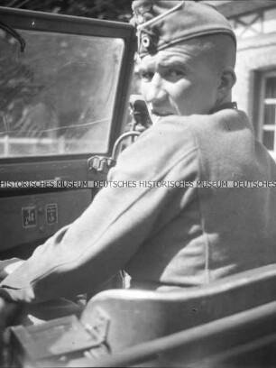 Ein deutscher Leutnant auf dem Beifahrersitz eines offenen PKW dreht sich zum Fotografen auf der Rückbank um