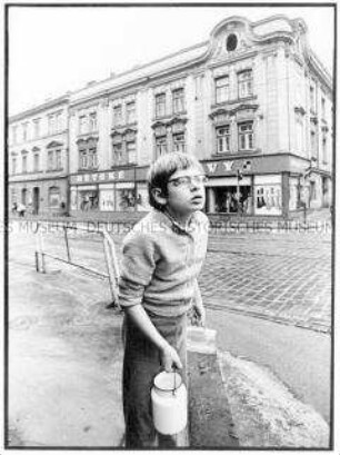 Junge am Straßenrand mit Milchkanne und Bierglas am Bordstein einer gepflasterten Straße in Prag stehend, im Hintergrund ein Geschäftshaus mit barocker Fassade (Altersgruppe 18-21)