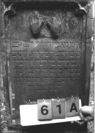 Grabstein 61 (Index: 99799529) Material: Sandstein Zustand: stark verwittert, versalzen Maße HxBxT: 118x62x22 cm Beschreibung: Stele, Abdeckplatte, Giebelaufsatz, Giebel abgeschlossen mit Palmette u. Voluten, Seitenakrotere, Schriftfeld eingetieft, Sockel Symbolik: segnende Hände: Grabstein 61A