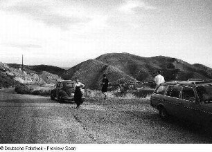 Zwei Autos und drei Personen in bergiger Landschaft mit See