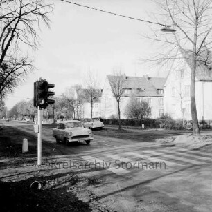 Sieker Landstraße: Fußgängerampel mit Zebrastreifen: links Pkw: dahinter Mitte parkender Pkw mit daneben stehender Person: hinten rechts drei Wohnblocks mit Grünanlage, Hecke und Bäumen