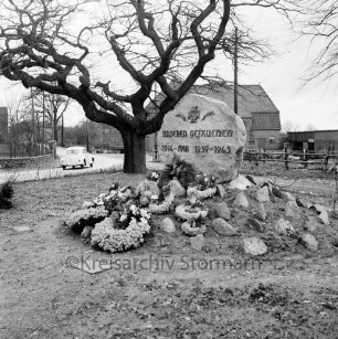 Ehrenmal mit Kranzschmuck und Baum: hinten links VW-Käfer: im Hintergrund Bauernhäuser
