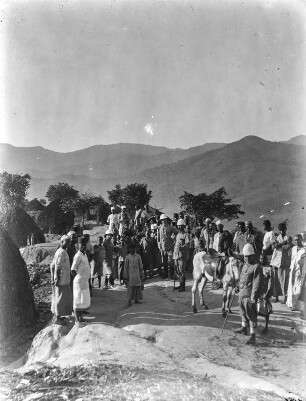 Dorfbewohner und Missionare : Eine Gruppe der Schambala und Missionare in Tropenanzügen vor dem Dorf Bumbuli auf einem Platz.