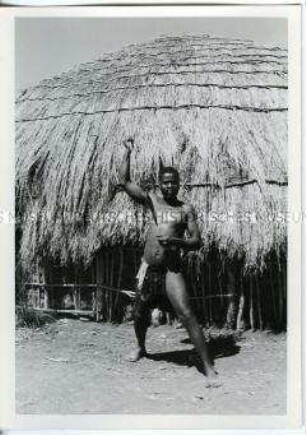 Mann der Volksgruppe Zulu mit erhobenem Speer vor einer Strohhütte, Südafrika