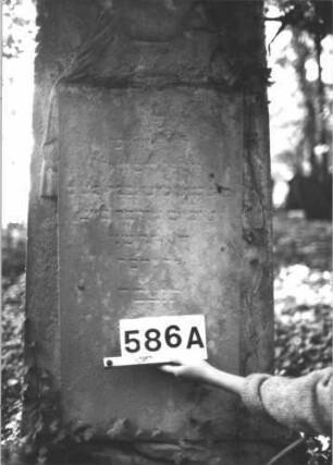 Grabstein 586 (Index: 99900727) Material: Sandstein Zustand: stark verwittert Maße HxBxT: 145x59x23 cm Beschreibung: aufgesockelte Stele mit Dreiecksabschluss, eingetiefter Rahmen, im Dreiecksfeld plastische Urne, mit Tuch bedeckt Symbolik: Urne, mit Tuch bedeckt: Grabstein 586A