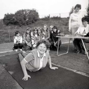 Liegestütze : Sportfest des Deutschen Turn- und Sportbundes (DTSB) in der DDR, zur Leibesertüchtigung der Bevölkerung. Kräftige Frau, um die 50 Jahre, versucht sich an den Liegestütze. Im Hintergrund sind weitere Frauen, von Jung bis Alt, zu sehen die sich über das Geschehen im Vordergund amüsieren beziehungsweise mitfiebern und bewerten