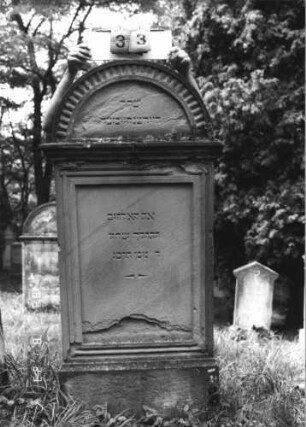 Grabstein 33 (Index: 100110895) Material: Sandstein Zustand: stark verwittert im Giebel, Abschieferungen im unteren Teil Maße HxBxT: 166x75x28 cm Beschreibung: Stele, Sockel, Rundbogenabschluß mit Zahnradfries und Verdachung, Schriftfelder eingetieft: Grabstein 33