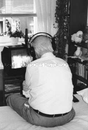 Älterer Mann sitzt mit Kopfhörern vor einem Fernseher (Altersgruppe 14-17)