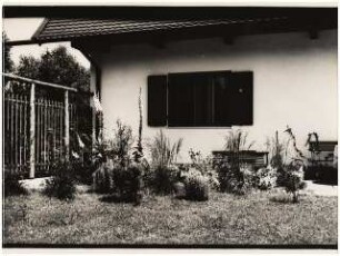 Wohnhaus und Garten [noch nicht indentifiziert]: Stauden (Eisenhut, Gräser) am Haus, dunkler Klappstuhl, gestreifter Liegestuhl, dunkle Fensterläden, Holzzaun mit Rankhilfe