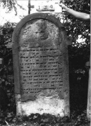 Grabstein 134 (Index: 100110701) Material: Sandstein Zustand: große Abschieferungen im Giebel und am unteren Rand, sonst leicht verwittert, geringer Textverlust Maße HxBxT: 145x84x15 cm Beschreibung: Stele, Rundbogenabschluß mit Ohren, Schriftfelder eingetieft: Grabstein 134