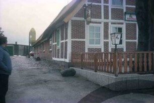 Papierholz 1: Gaststätte Zur Hahnheide: giebelständiges Bauernhaus mit Reetdach: Teilansicht: Straßenfront mit Fachwerk und Wirtshausschild, davor Terrasse mit Geländer, Laterne, alter Baum: Traufseite: Hof, hinten links Wirtschaftsgebäude, Silo: im Vordergrund links Mann