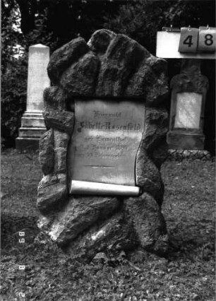 Grabstein 48 (Index: 100101473) Material: Kalkstein, weiße Marmorplatte Zustand: gut Maße HxBxT: 117x57x37 cm Beschreibung: Stele, rustica, Schriftplatte in Pergamentrollenform Steinmetz: J. Götz: Grabstein 48