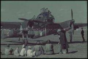 Einheimische vor einem Flugzeug der deutschen Wehrmacht sitzend