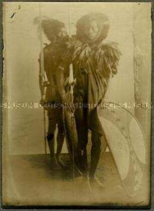 Zwei hintereinander stehende Massai-Krieger in Schmuck und Waffen