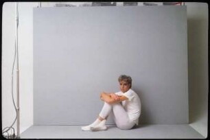 Fotoshooting einer Performance- oder Schauspielgruppe : Ein Schauspieler in weißem Kostüm vor einem grauen Hintergrund sitzend