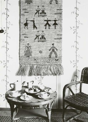 "Gardinen und Tapeten". Interieur mit Korbsessel und Korbtisch vor Tapete mit Blattmuster und Wandteppich mit Figuren