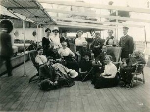 Gruppenbildnis von stehenden und sitzenden Passagieren an Deck eines Hochseepassagierdampfers
