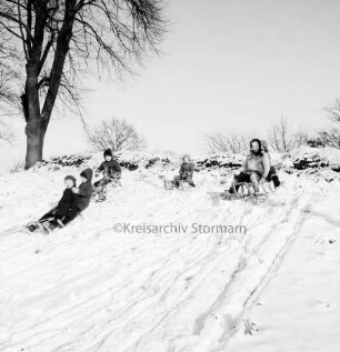 Dorfstraße: Kinder mit Schlitten beim Rodeln an Straßenböschung: links Baum: Winter
