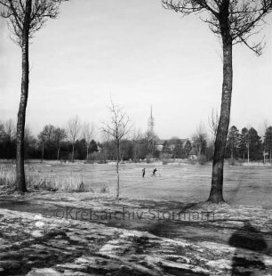 Groten Diek: Eisdecke: in der Mitte zwei Kinder, eines mit Fahrrad auf Teich: vorn Weg, rechts und links drei Bäume und Schilf: am gegenüberliegenden Ufer Turm der Auferstehungskirche, Häuser und Bäume: Winter