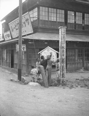 Wegweiser (wahrscheinlich in Sapporo) : Straßenbild mit Mann und Jungengruppe, einen Aushang neben einem Gasthaus anbringend