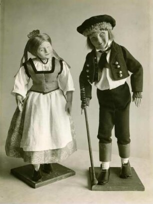 Paar aus Landau. Zwei handgefertigte Spielfiguren aus Holz, in traditioneller Stoffbekleidung. Frau mit Schürze und Kleid, Mann mit Hut, und Gehstock. Halbtotale