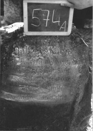 Grabstein 574 (Index: 100111470) Material: Granit (?) Zustand: Platte mehrfach gesprungen mit Textverlust, sonst gut Maße HxBxT: 105x94x40 cm Beschreibung: Stele, rustica, Segmentbogenabschluß, ovale Schriftplatte: Grabstein 574A