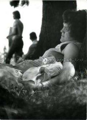Eine Frau liegt auf dem Rasen, an einen Baumstamm gelehnt, in ihrem Arm liegt ein Baby
