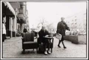 Ältere Dame mit Tasche sitzt in einem Sessel auf dem Bürgersteig