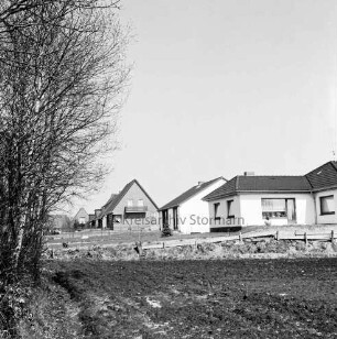 Neubausiedlung mit Einfamilienhäusern: links Bäume: vorn Feld, dahinter Bauzaun