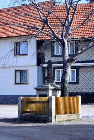 Gedenkstein und Laubbaum, dahinter Häuser