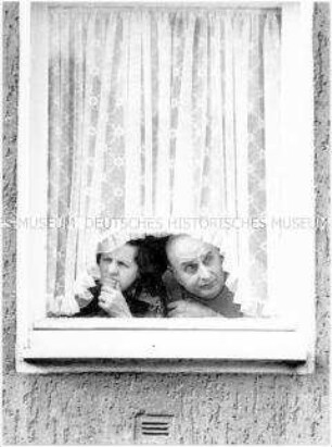 Ehepaar schaut unter der Gardine aus dem Fenster heraus (Altersgruppe 18-21)