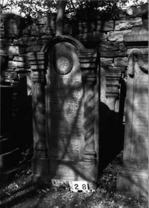 Grabstein 28 (Index: 99629529) Material: Sandstein Zustand: verwittert/leichter Textverlust Maße HxBxT: 152x69x10 cm Beschreibung: Stele mit eingezogenem Rundbogenabschluß mit Verdachung, zwei gedrehte Halbsäulen Symbolik: gedrehte Halbsäulen, Kranz: Grabstein 28