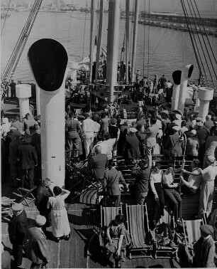 Ein Passagierschiff läuft in einen Hafen ein. Passagiere stehen an Deck.