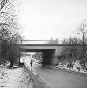 Landesstraße zwischen Reinfeld und Westerau: hinten Mitte Fahrradfahrer: dahinter Brücke der Autobahn A 1: rechts und links Böschung mit Büschen und Bäumen: Winter