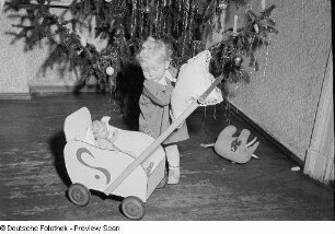 Porträt eines kleinen Mädchens mit Puppenwagen vor einem Weihnachtsbaum