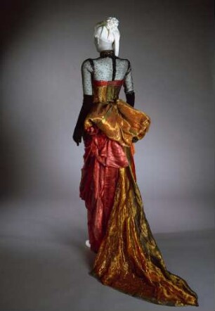 Hochgeschlitztes Auftrittskleid aus rot-goldenem Lamé mit aufwändigen Drapierungen, dazu eine transparente Bluse mit Jetperlenbesatz und eine Reitpeitsche (Archivtitel)