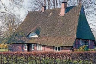 Kamp 12: Fachwerkkate mit Reetdack: Traufseite, links Wohntrakt mit Eingangstür, Mitte Sprung, rechts Wirtschaftstrakt: Giebelseite ehemaliger Wirtschaftstrakt, Scheunentor mit Verglasung: vorne Gartenhecke