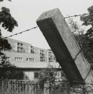 Zaunsäule mit Stacheldraht vom Maschendrahtzaun (ehem. Grenze des Geländes der Staatssicherheit) des Gebäudekomplexes Nr. 112 - 122, dahinter eine Baracke, ein Nebengebäude und die östlich angrenzende 5-geschossige Wohnbebauung