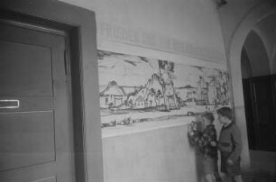 Dekorative Ausgestaltung der Klassenräume und Schulflure in der Einheitsschule Baumschulenweg und Oberschule Niederschöneweide in Vorbereitung des Deutschlandtreffens der Jugend für Frieden und Völkerfreundschaft zu Pfingsten 1950 : Schüler beim Anbringen eines Plakats zum Deutschlandtreffen an einer Wand im Hausflur einer Schule