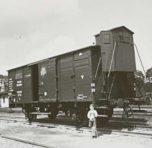 Zweiachsiger gedeckter Güterwagen (Gattungsbezirk Karslruhe) der Deutschen Reichsbahn mit Flachdach und Bremserhaus auf einem Abstellgleis, davor posierender Junge