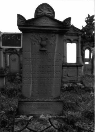 Grabstein 1204 (Index: 100104371) Material: Sandstein Zustand: am Sockel und im Giebelbereich stark verwittert Maße HxBxT: 164x48x15 cm Beschreibung: Stele auf getrepptem Sockel, Giebelausatz mit Giebel- und Eckakroterien, gefüllt mit Blütenornamenten, unterhalb des Giebels plastische, aufgesetzte Levitenkanne, unterster Sockel felsartig gearbeitet Symbolik: Levitenkanne, Blüten: Grabstein 1204