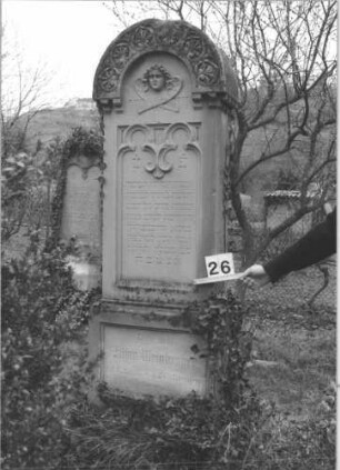 Grabstein 26 (Index: 99805082) Material: Sandstein Zustand: leicht verwittert Maße HxBxT: 207x58x26 cm Beschreibung: Stele auf Doppelsockel mit Rundbogen, Inschriftennische rechteckig mit zwei Kleeblattbögen als Verzierung, Inschriftennische im Sockel querrechteckig Symbolik: Engel im Rundbogen, Blattornamentik am Rundbogen Besonderheit: kulturhistorisch wertvoll / künstl.-formale Gründe: Grabstein 26