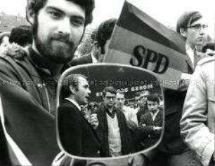 Autospiegel, in dem sich ein mit Mikrophon sprechender SPD-Kandidat mit Zuhörern spiegelt (Altersgruppe 18-21)