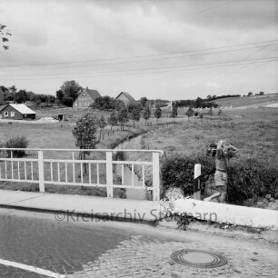 Norderbeste nach Regulierung: hinten Mitte Baumreihe am Ufer: rechts und links Wiesen: hinten links Neubau eines Hauses: hinten Mitte Bauernhäuser: Blick von der Brücke der Bundesstraße B 75 mit alter Straßenführung auf das Unterdorf: rechts Kind mit Tasche neben Geländer