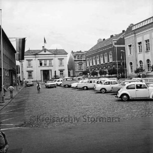 Markt: vorne parkende Pkws, links die Kreissparkasse mit Beflaggung, im Hintergrund links das Rathaus, rechts daneben das Stadthaus: Blick von Südosten