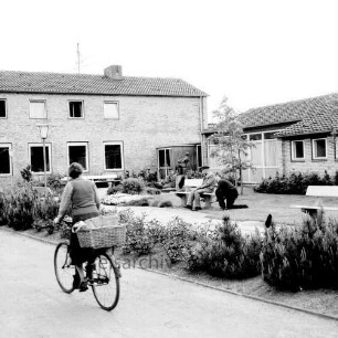 Ahrensburg: Reeshoop: Außenaufnahme: links Wirtschaftsgebäude, rechts Wohnflügel: Grünanlage mit Parkbänken, Parklampe, jungen Bäumen, Sträuchern: Heimbewohner auf Bank, Arbeiter bei Rasenpflege: Kunst am Bau, Bronzeplastik "Ziehharmonikaspieler" von Bildhauer Karlheinz Goedtke: vorne links Zufahrt, Fahrradfahrerin
