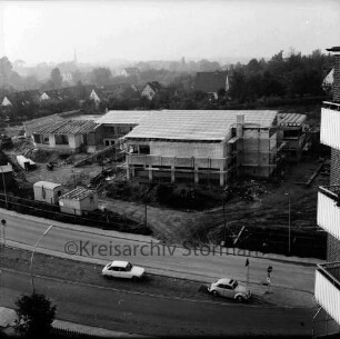 Evangelisches Gemeindehaus: Masurenweg: Rohbau: Blick von einem Hochhaus auf die Baustelle mit Bauwagen, Erdhaufen, Bretterstapel, Förderband und Baumaterial: vorn zwei Pkw auf Straße: hinten Einfamilienhäuser, Gärten und Bäume: im Hintergrund links Turm der Peter-Paul-Kirche im Dunst