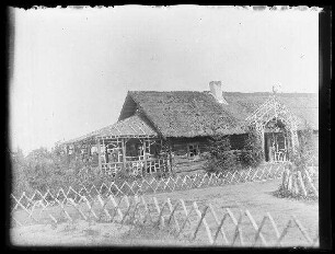 Unbekannte Ortschaft [Litauen/Weißrussland/Ukraine?]: reetgedecktes Holzhaus mit Birkenholzveranda, Birkenholzeingang, im Vordergrund ein Jägerzaun aus Birkenholz (Nahaufnahme)