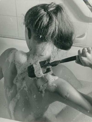 In der Badewanne. Eine Frau massiert sich mit einer Bürste ihre rechte Schulter. Badeschaum befindet sich auf ihrer Haut